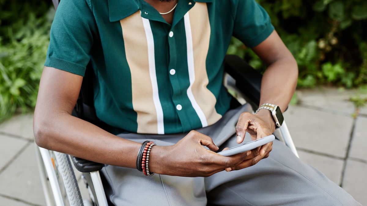 Person in a wheelchair wearing a green and tan striped shirt, gray pants, and a smartwatch, using a smartphone outdoors.