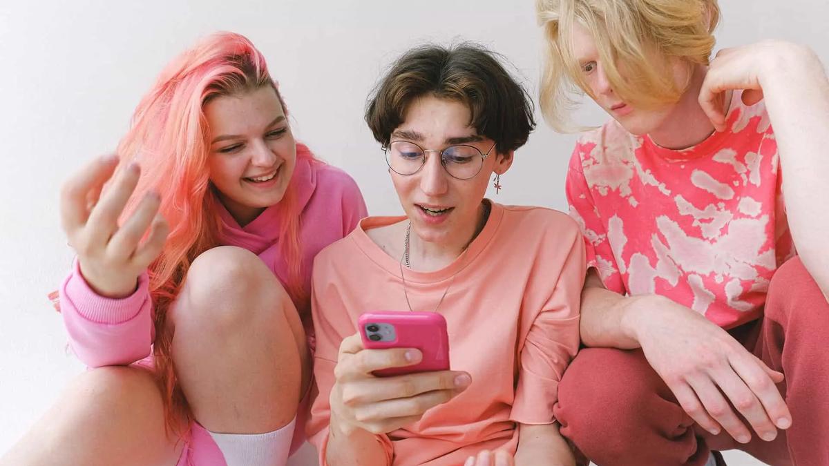 Three young people in pink and red clothing looking at a pink smartphone, white background, happy mood.