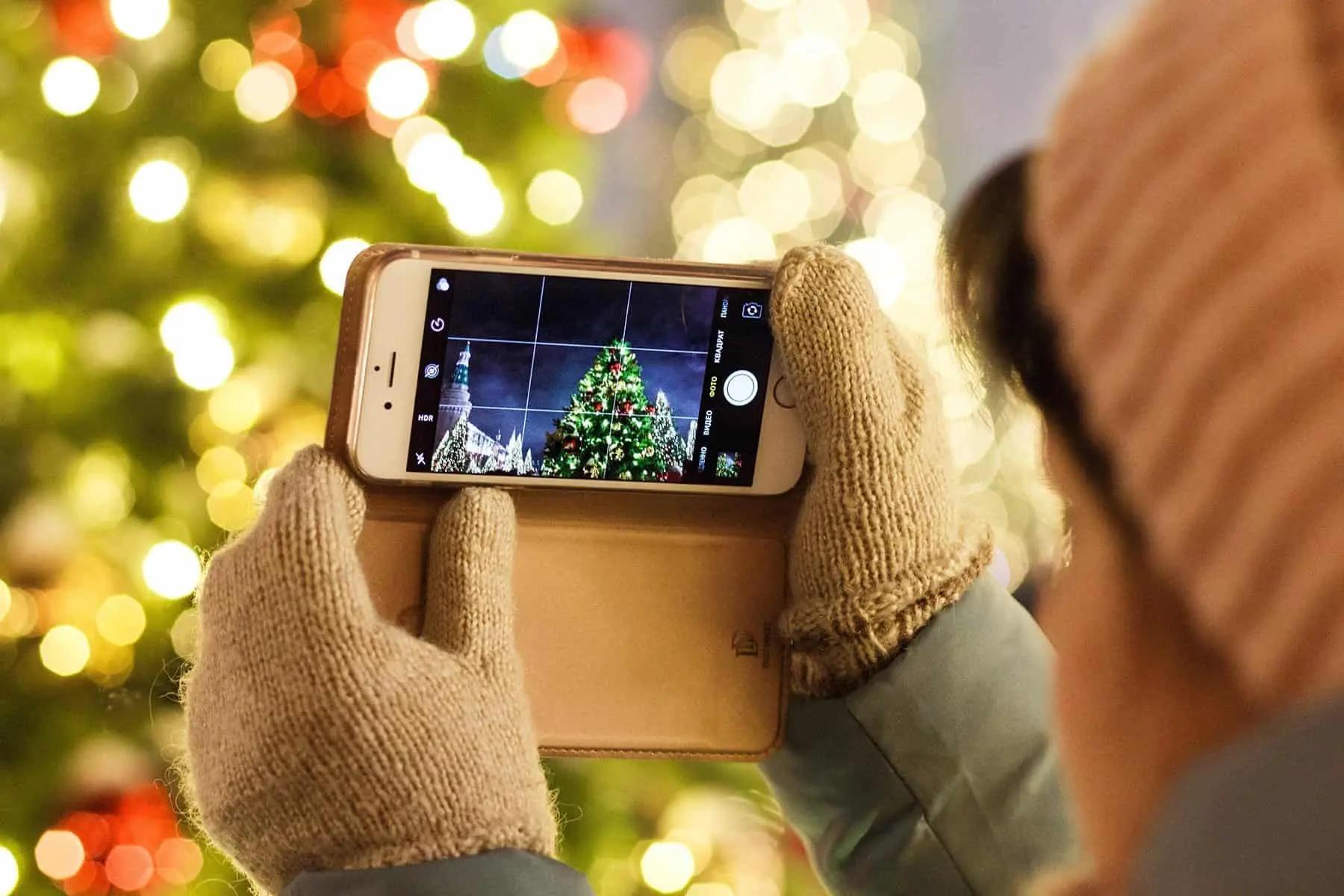 Una persona con guantes de lana fotografía un árbol de Navidad iluminado con la cámara de su móvil