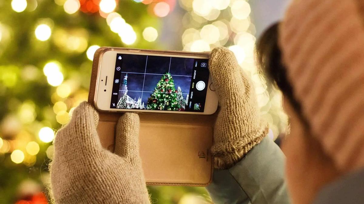A person in mittens photographs a lit Christmas tree using a smartphone camera app