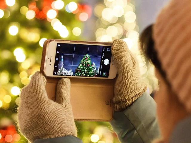 A person in mittens photographs a lit Christmas tree using a smartphone camera app