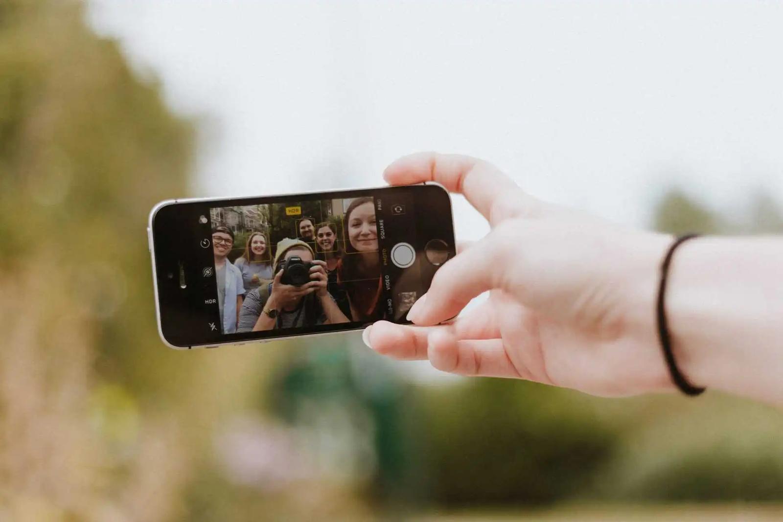A hand holds up a phone taking a photo of a group of friends posing outdoors