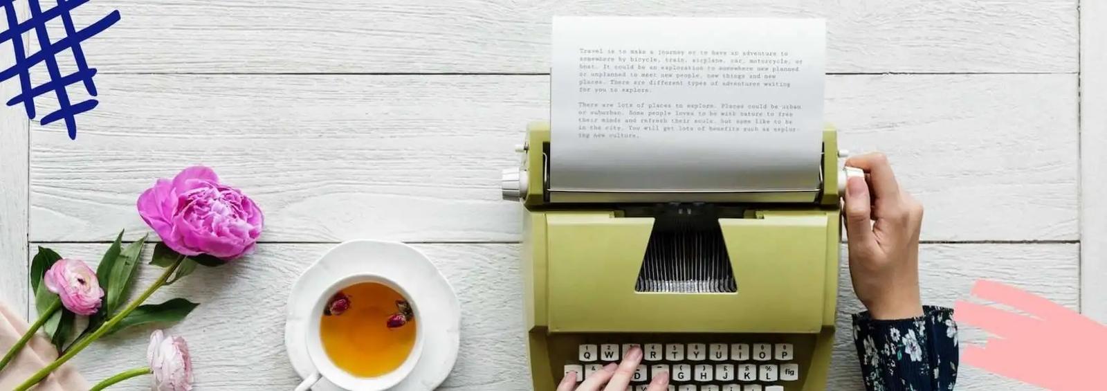 Hands typing on a vintage typewriter beside a cup of tea and pink flowers on a wooden table