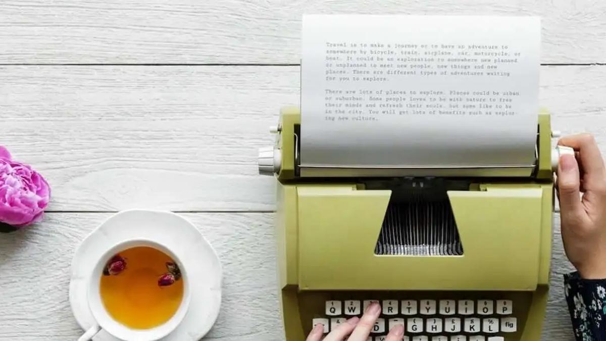 Hands typing on a vintage typewriter beside a cup of tea and pink flowers on a wooden table