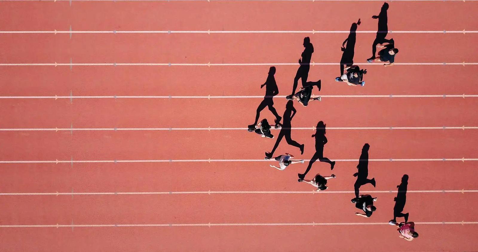 Vista aerea de corredores compitiendo en una pista de atletismo roja, proyectando largas sombras