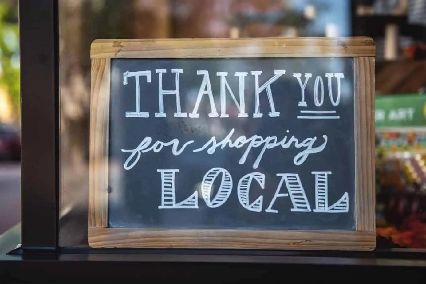 A wooden-framed chalkboard sign in a shop window reading "Thank you for shopping local"