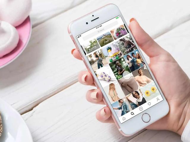 A hand holds a phone showing the Instagram explore grid with shopping and travel category tags