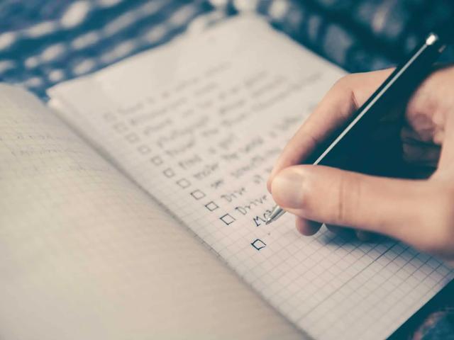 A hand holding a black pen, writing on a checklist in a notebook. The text includes 'Drive' and 'Me'.