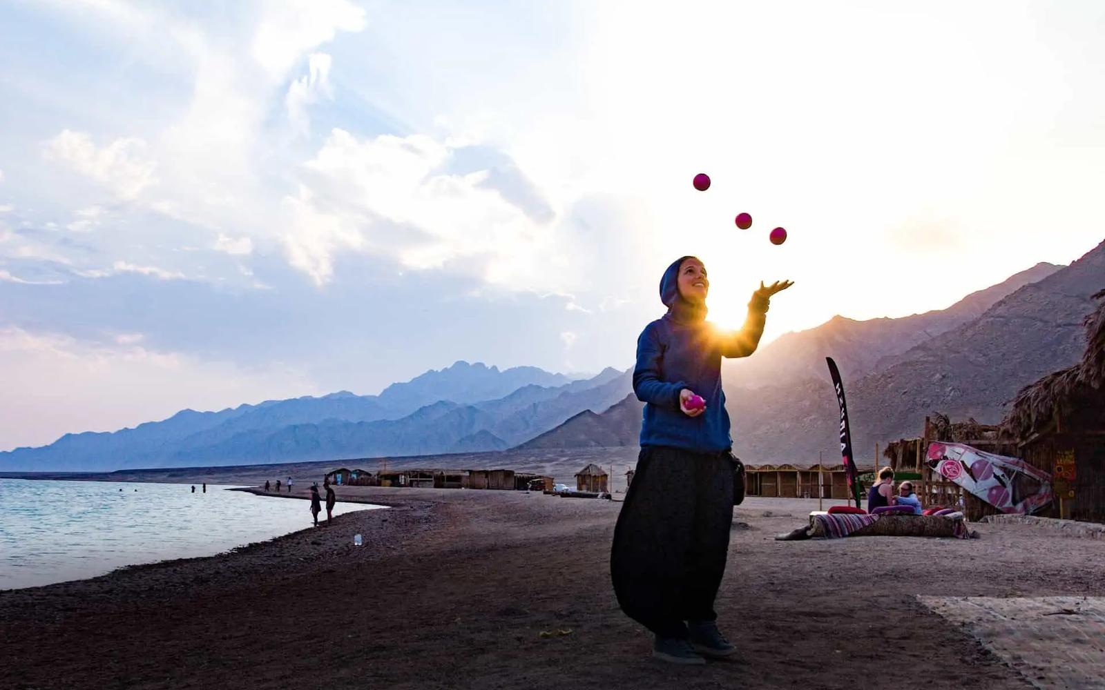 A person juggling pink balls on a beach at sunset with mountains in the background