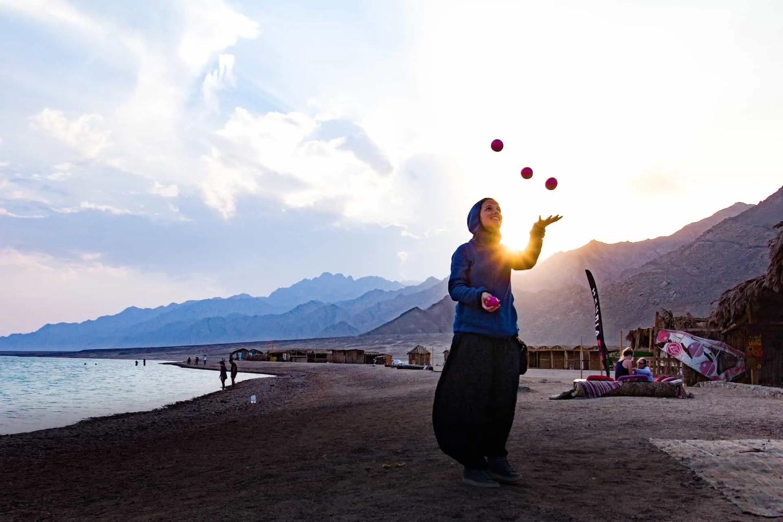 A person juggling pink balls on a beach at sunset with mountains in the background