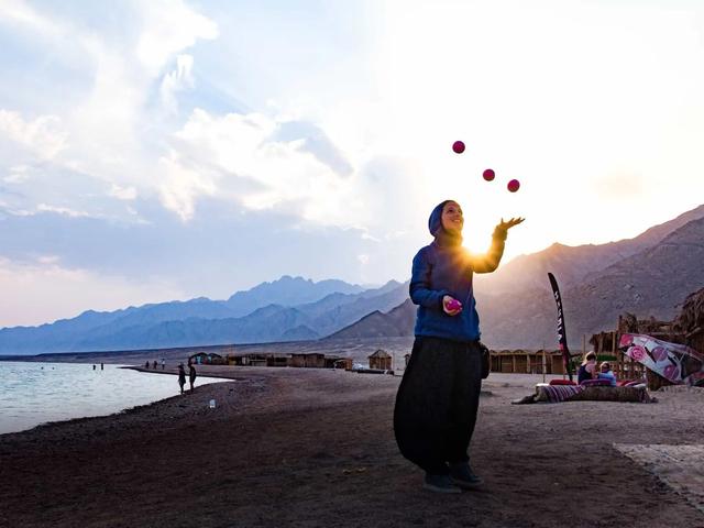 Una persona hace malabares con pelotas rosas en una playa al atardecer, con montañas al fondo