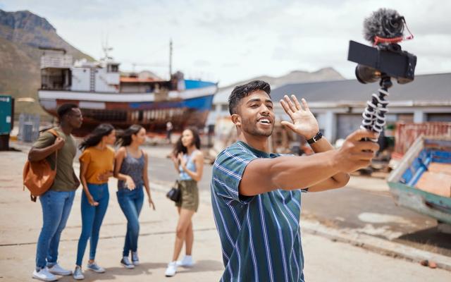 Man holding a camera on a tripod, waving, with four people and a large boat in the background, daytime, outdoor setting