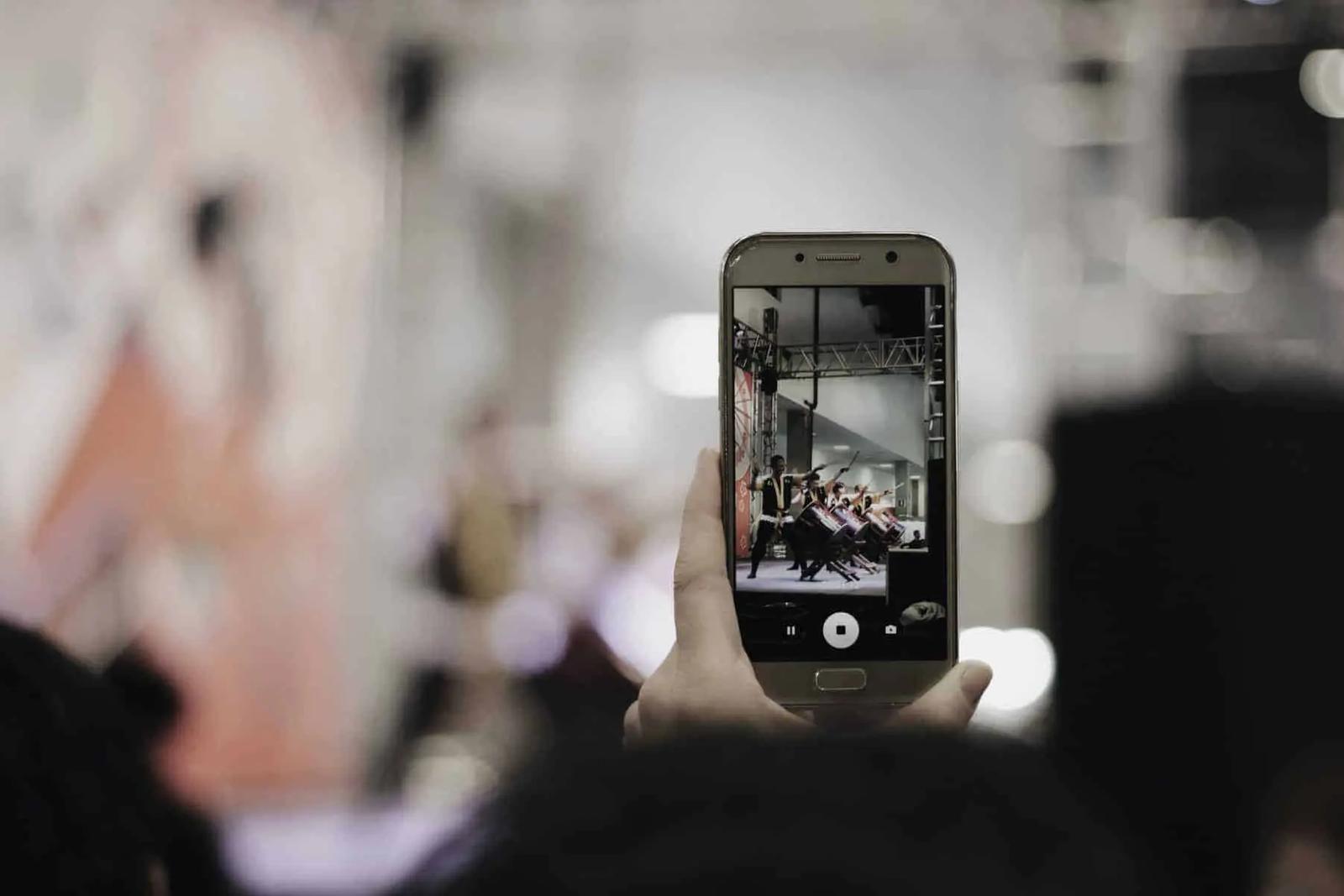 Una mano sostiene un teléfono grabando en video a artistas sobre un escenario