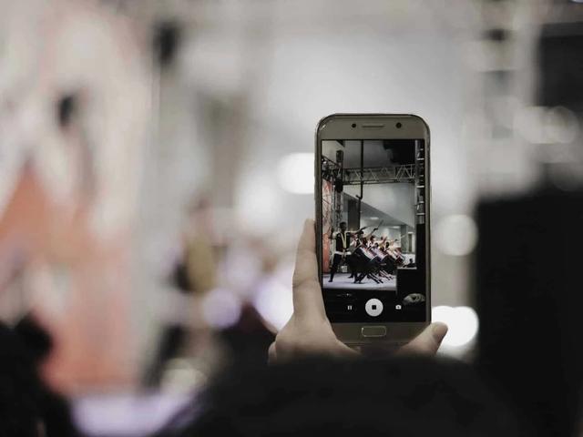 A hand holds up a smartphone recording video of performers on a stage