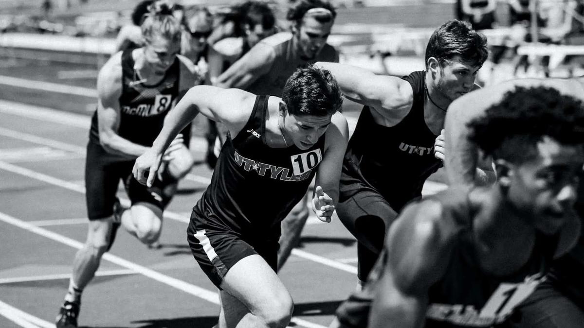 Runners in team jerseys sprinting off the start line on a track in a black and white photo