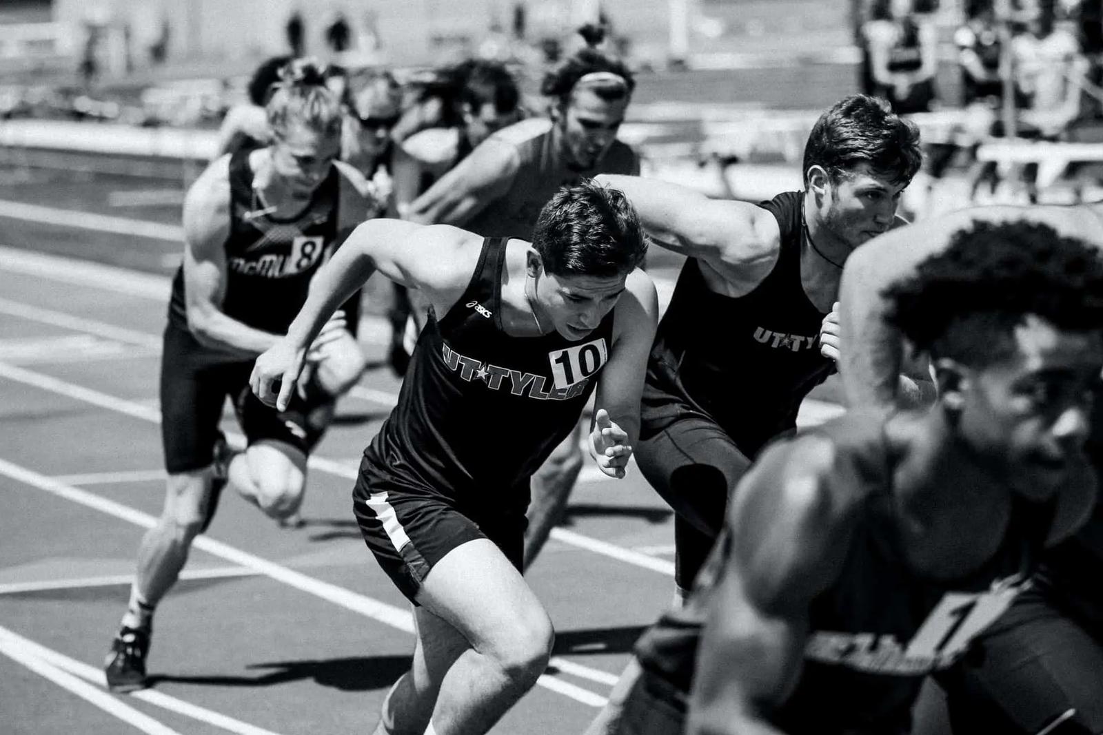 Runners in team jerseys sprinting off the start line on a track in a black and white photo