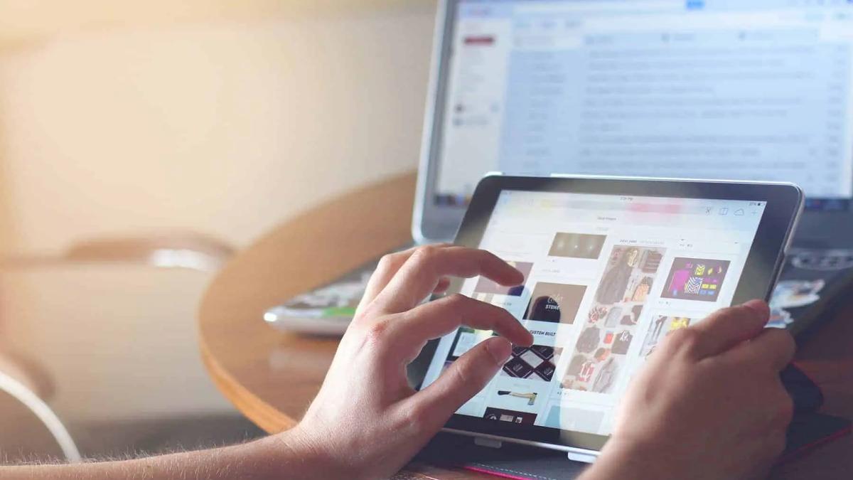Hands holding and scrolling a tablet displaying a grid of images, with a laptop in the background. Warm light.