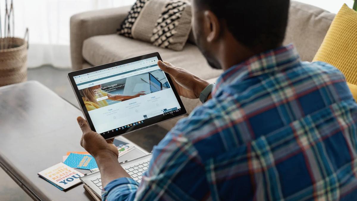 Man in blue plaid shirt holding a tablet displaying the LinkedIn Sales Navigator website, laptop on table, sofa in background