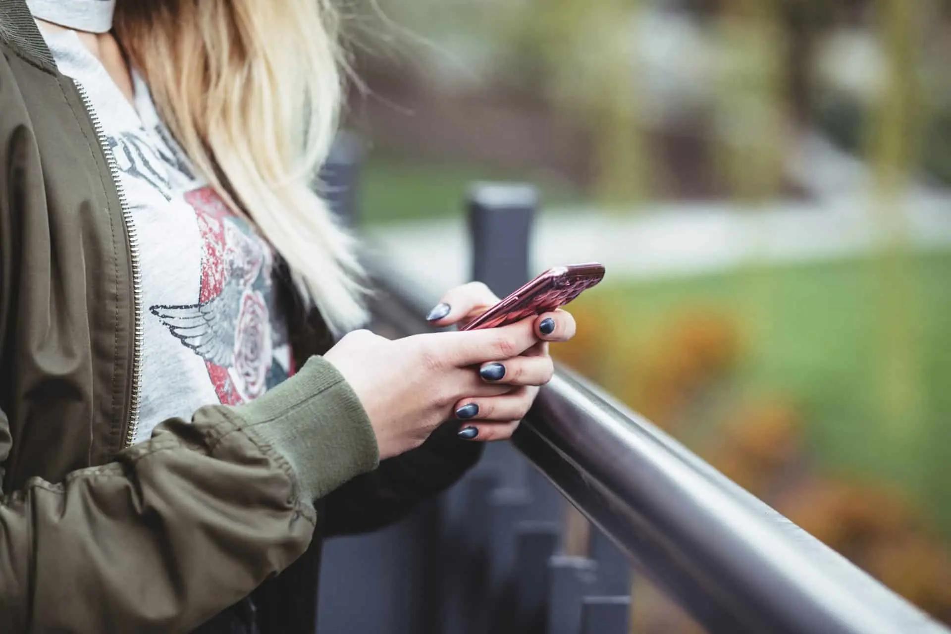 A woman with blonde hair leans on a railing outdoors while checking her phone.