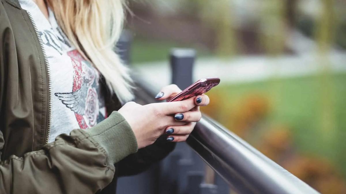 A woman with blonde hair leans on a railing outdoors while checking her phone.
