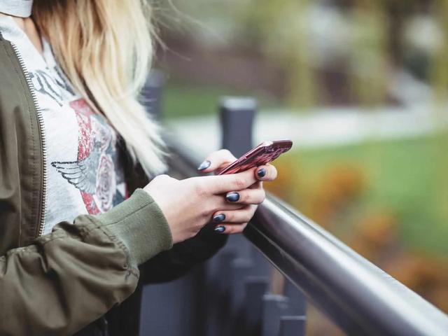 Una mujer rubia apoyada en una baranda al aire libre mientras revisa su teléfono.