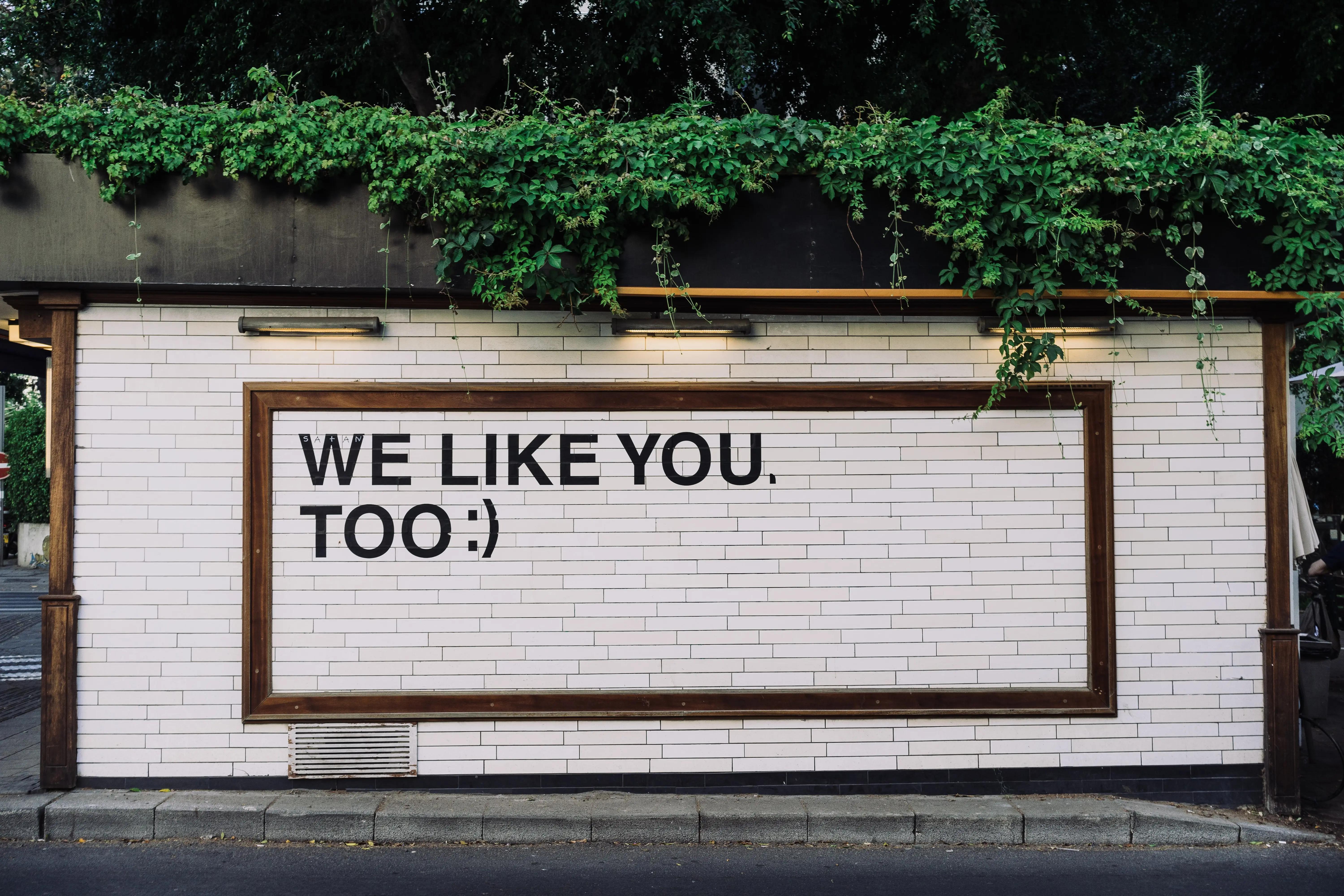 Un letrero enmarcado en una pared de baldosas que dice 'We like you, too :)' bajo enredaderas verdes colgantes