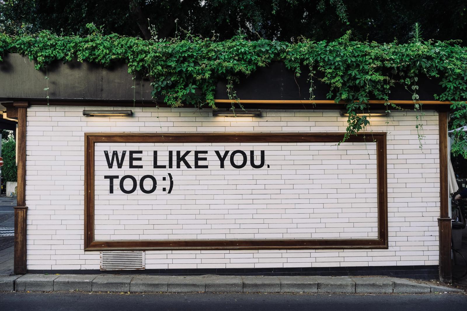 A framed sign on a tiled wall reading 'We like you, too :)' beneath overhanging green vines