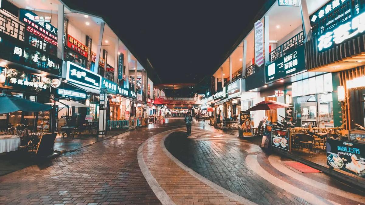 Una calle peatonal empedrada de noche, bordeada de tiendas iluminadas con letreros de neón