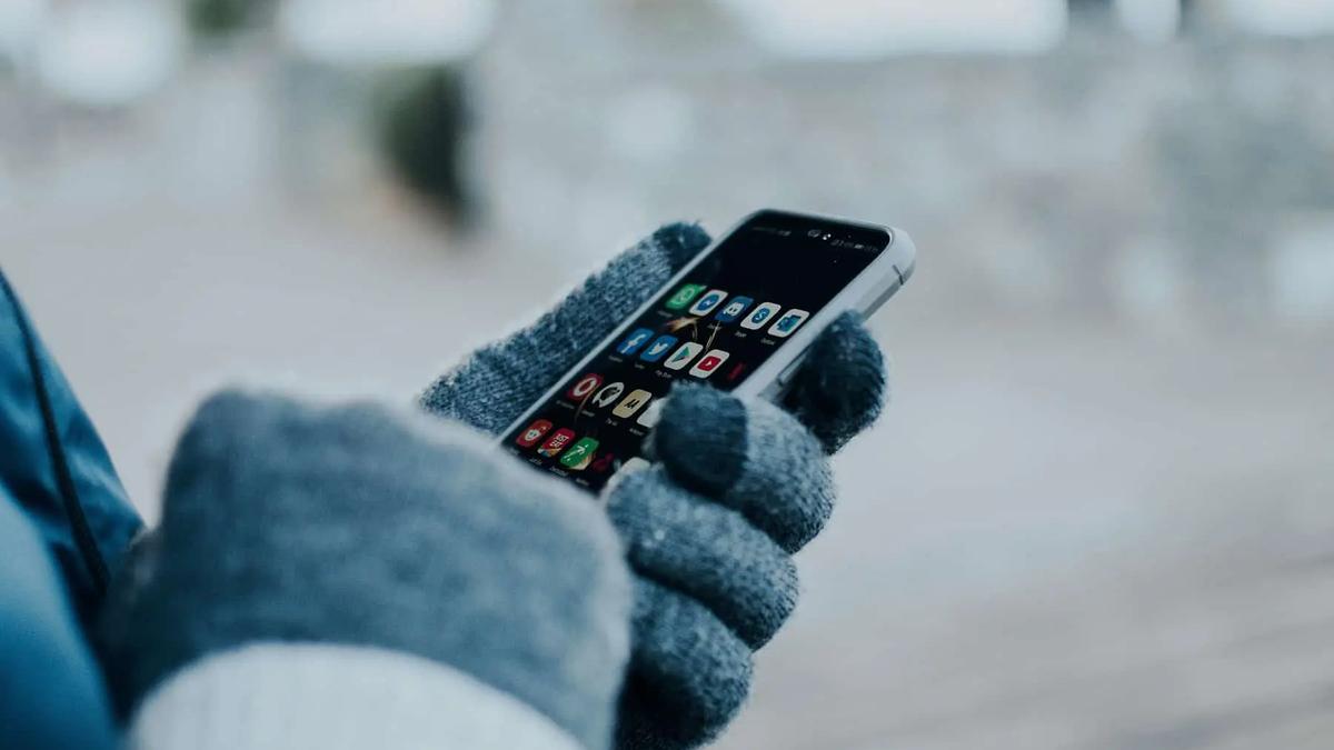 Hands in gray gloves holding a white smartphone with various app icons on a dark screen, blurred background.