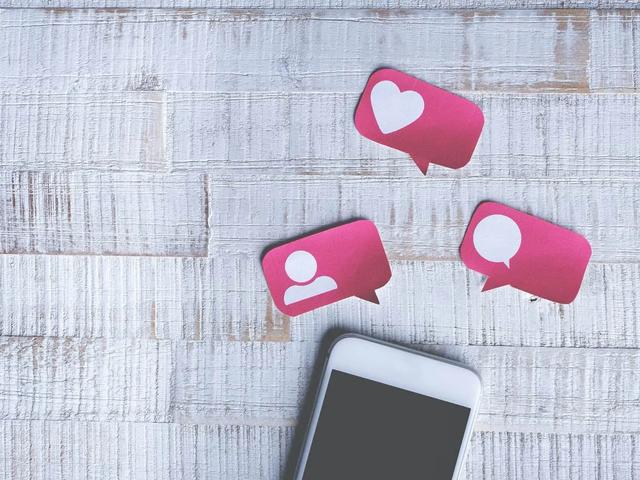 White smartphone with dark screen, three pink social media notification icons with heart, person, and speech bubble symbols, on a white wooden surface. Flat