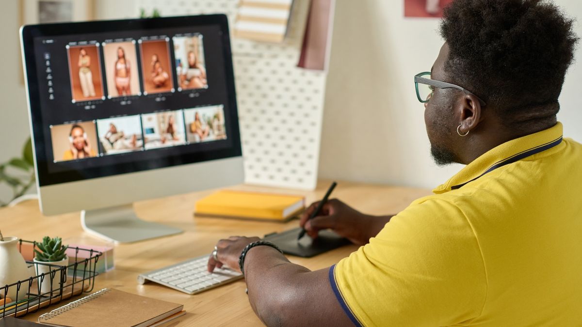 Man in yellow shirt and glasses editing photos on a desktop computer with a pen tablet, light wood desk. Neutral mood.