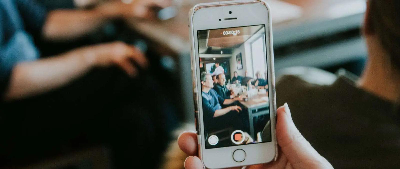 A hand holds up a phone recording video of people seated at a restaurant table