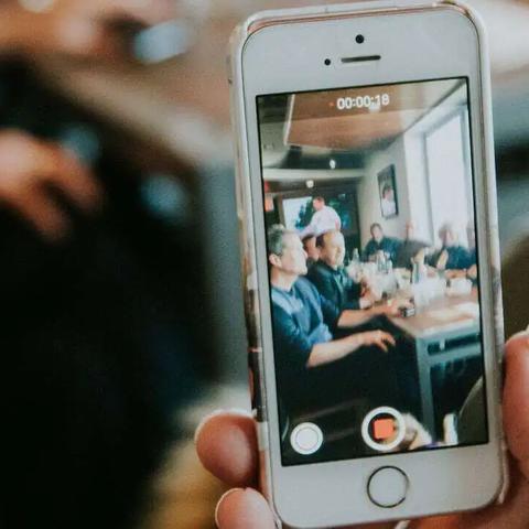 A hand holds up a phone recording video of people seated at a restaurant table