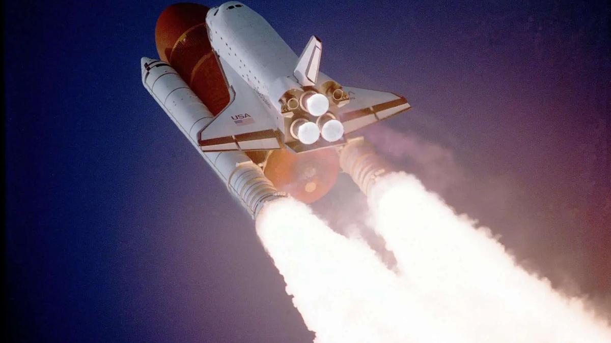 Space shuttle launching into a dark blue sky, with bright white and orange exhaust plumes from its engines. Text reads USA.