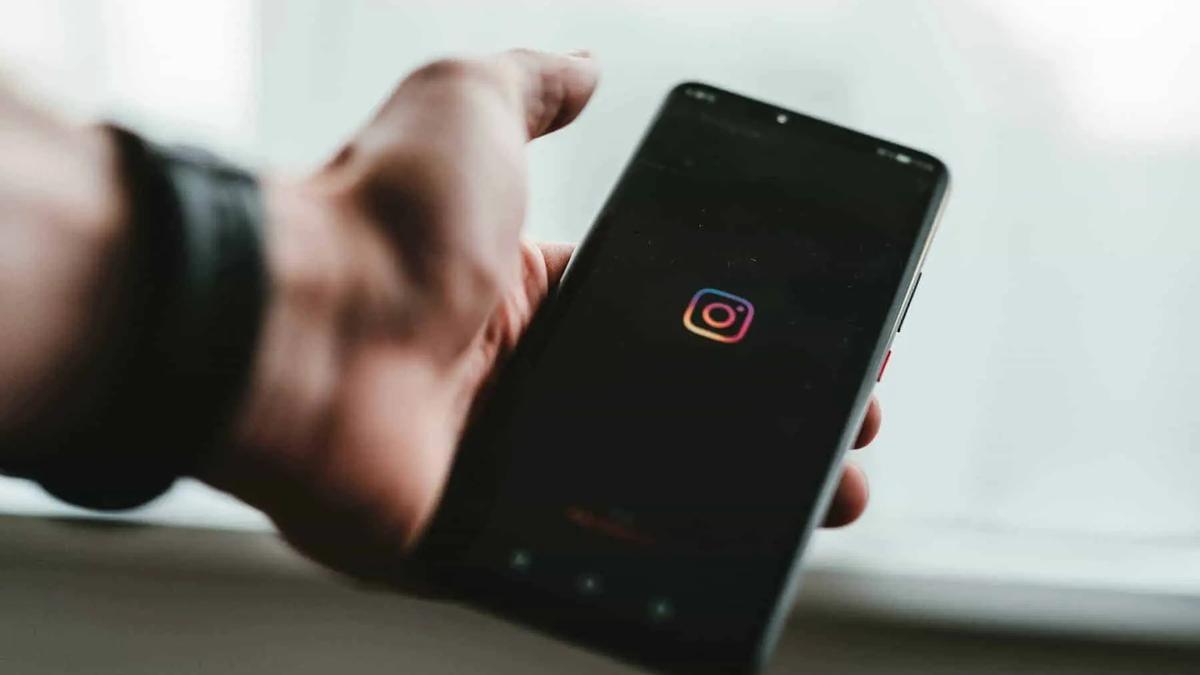 Hand holding a black smartphone with the Instagram app icon on the screen. The background is blurred and light.
