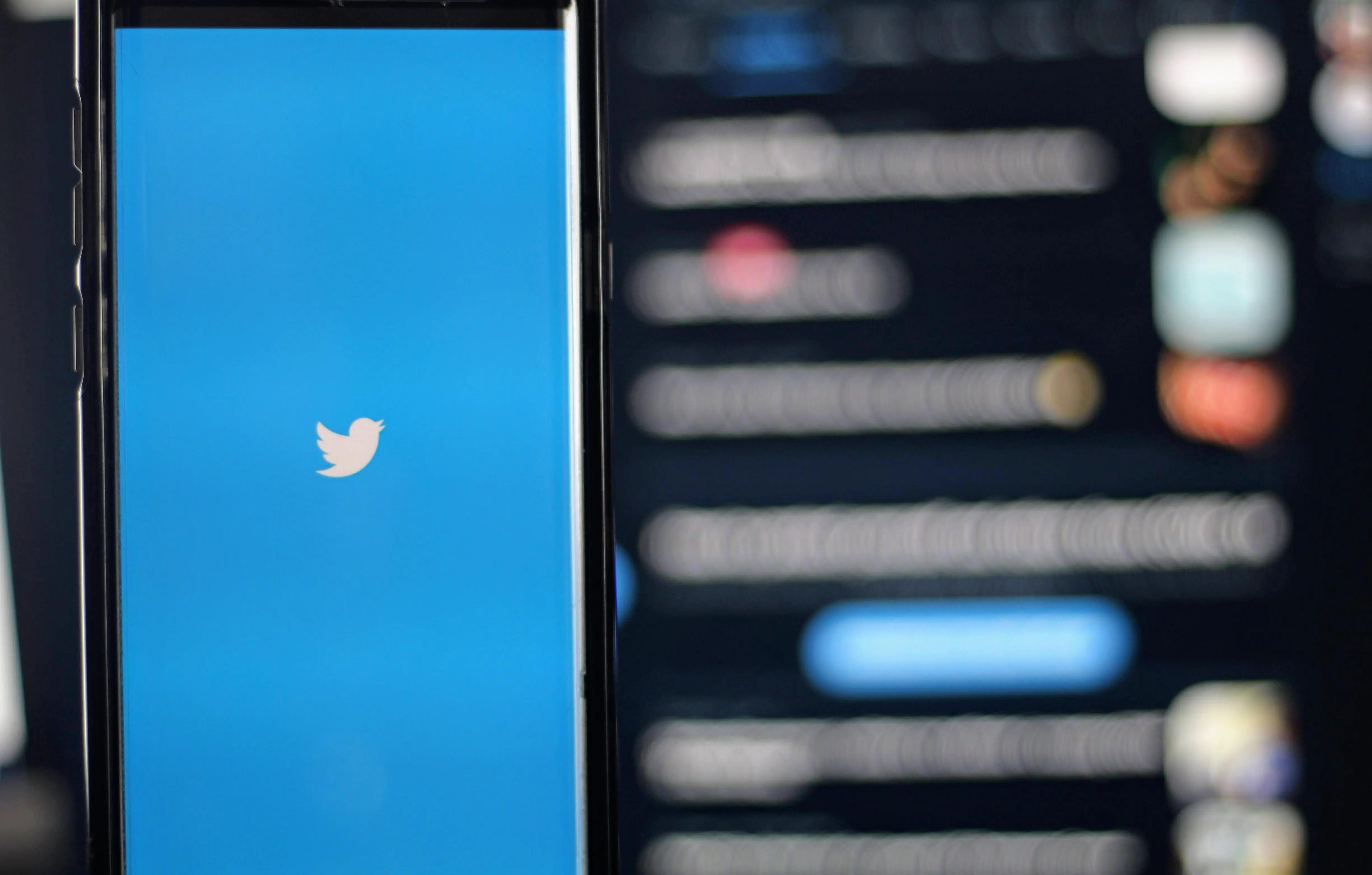 A smartphone showing the Twitter bird logo on a blue screen, with a blurred feed in the background.