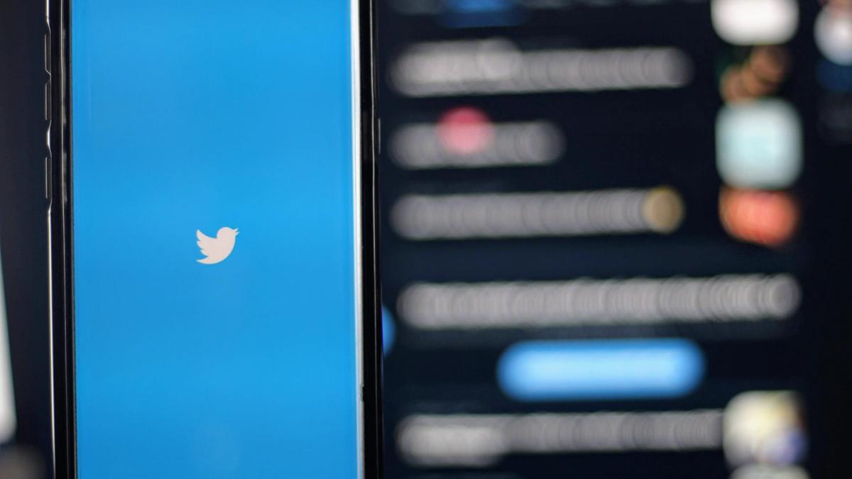 A smartphone showing the Twitter bird logo on a blue screen, with a blurred feed in the background.