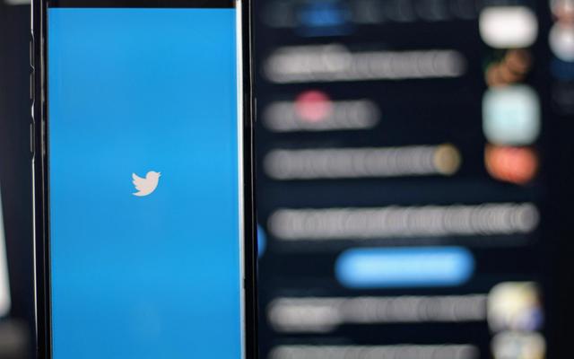 A smartphone showing the Twitter bird logo on a blue screen, with a blurred feed in the background.