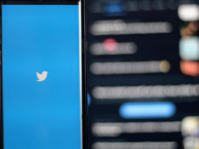 Un smartphone con el logo del pájaro de Twitter en pantalla azul, con un feed desenfocado de fondo.