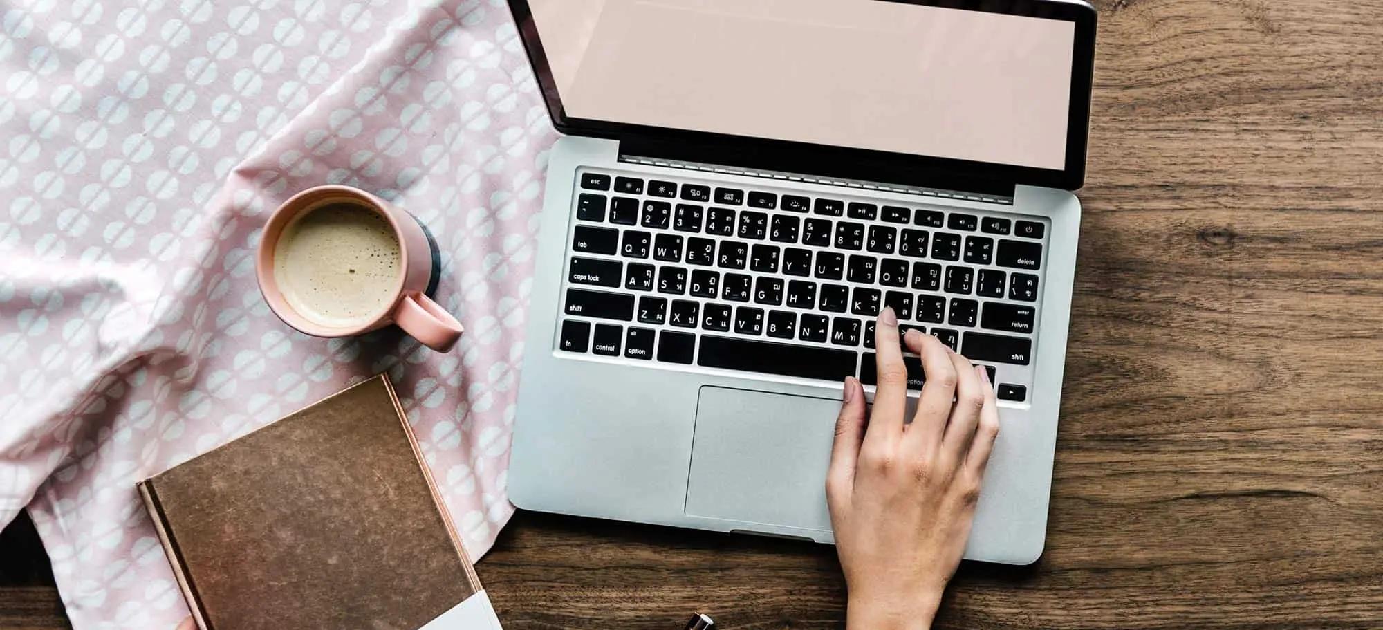 Overhead view of a hand typing on a laptop beside a coffee cup and notebook on a wooden desk