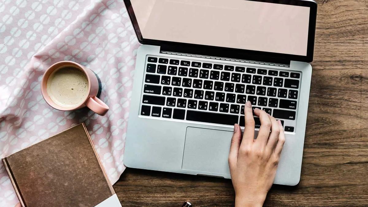 Overhead view of a hand typing on a laptop beside a coffee cup and notebook on a wooden desk