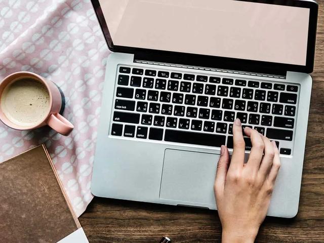 Vista cenital de una mano escribiendo en un portatil junto a una taza de cafe y un cuaderno sobre un escritorio de madera