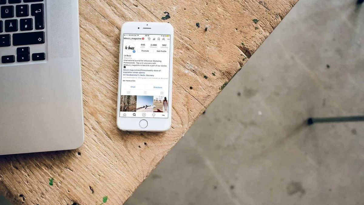 White iPhone displaying an Instagram profile, next to a silver laptop on a wooden and concrete surface