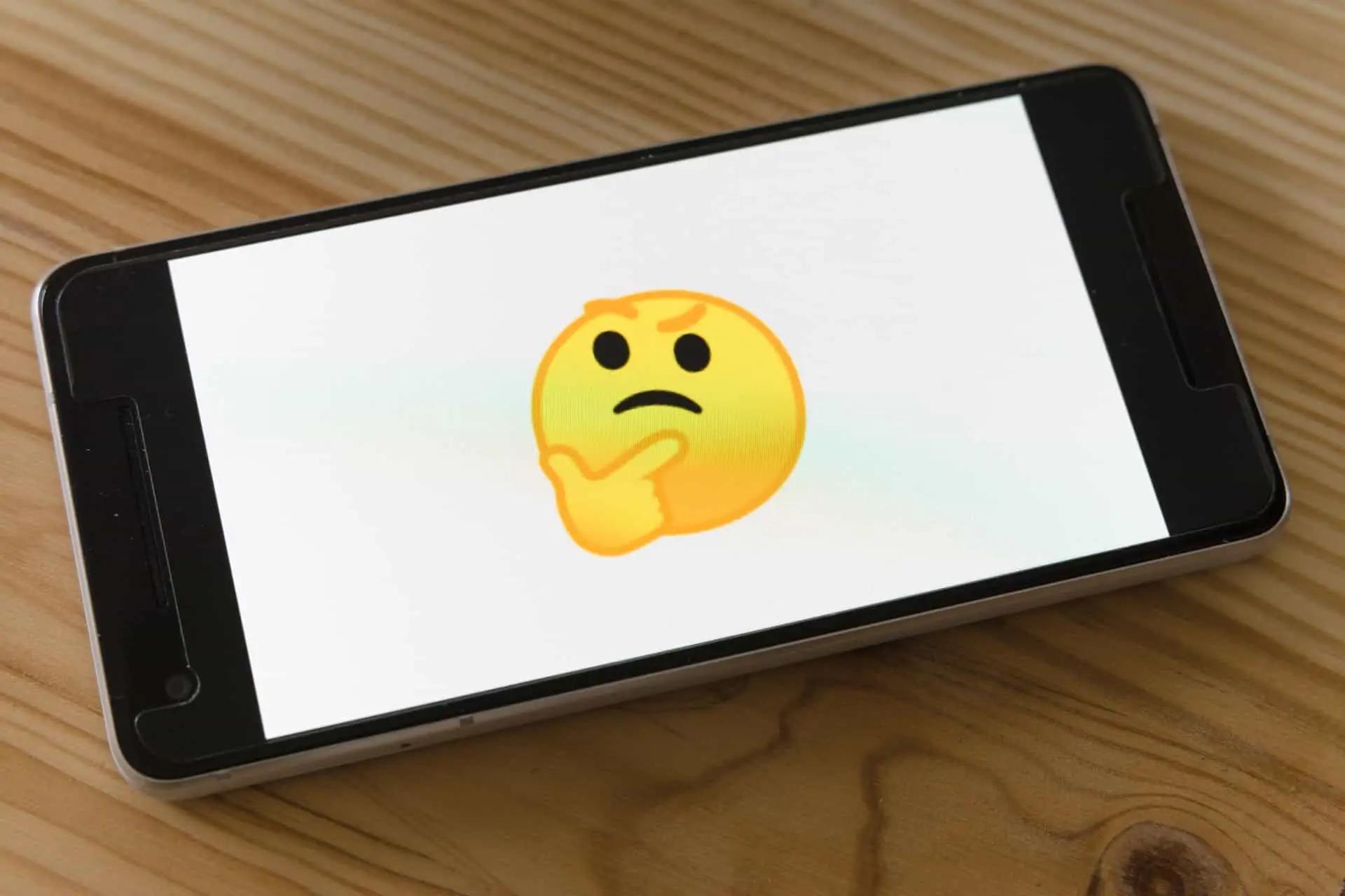 A smartphone lying on a wooden table displaying a large thinking-face emoji on screen