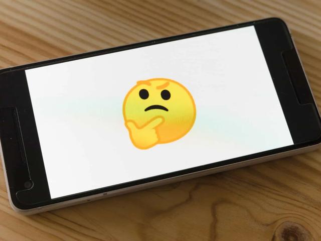 A smartphone lying on a wooden table displaying a large thinking-face emoji on screen