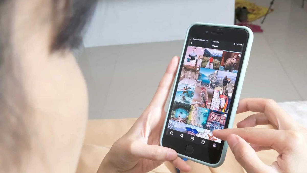 Person holding a black smartphone with a light blue case, browsing Instagram's travel photos. UI shows 'Travel' text.