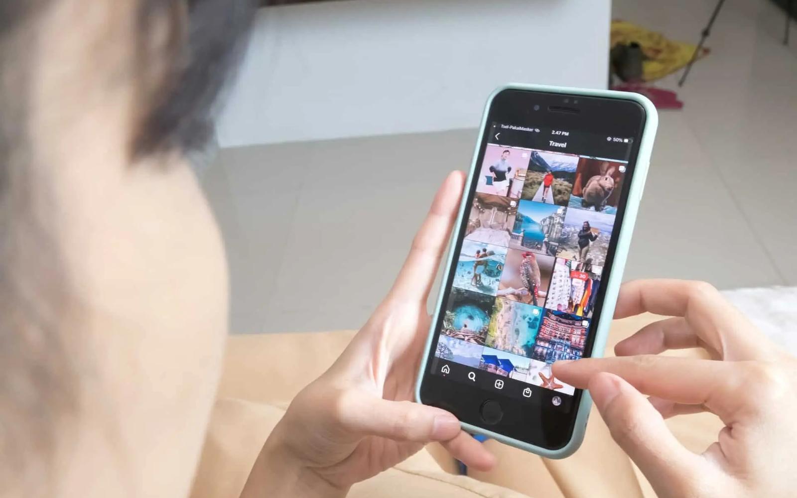 Person holding a black smartphone with a light blue case, browsing Instagram's travel photos. UI shows 'Travel' text.