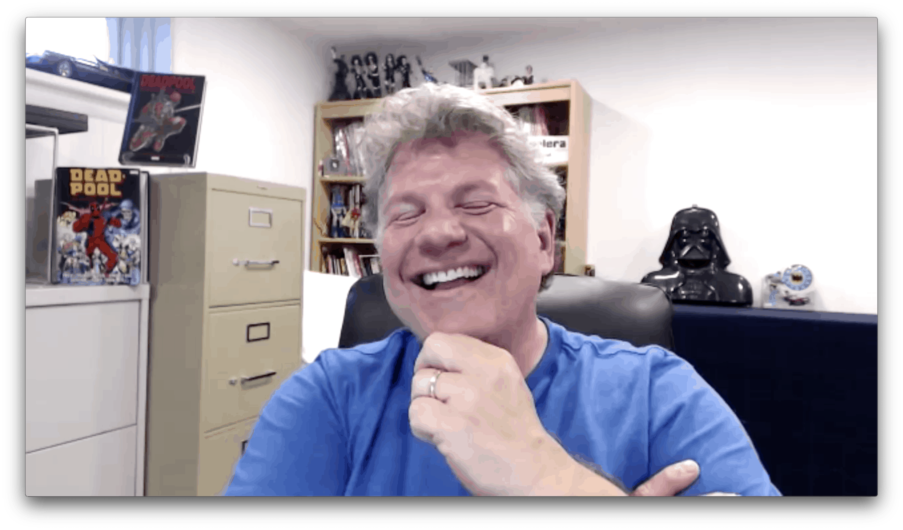 Buddy Scalera riendo en una videollamada, con arte de cómics y figuras de colección al fondo.