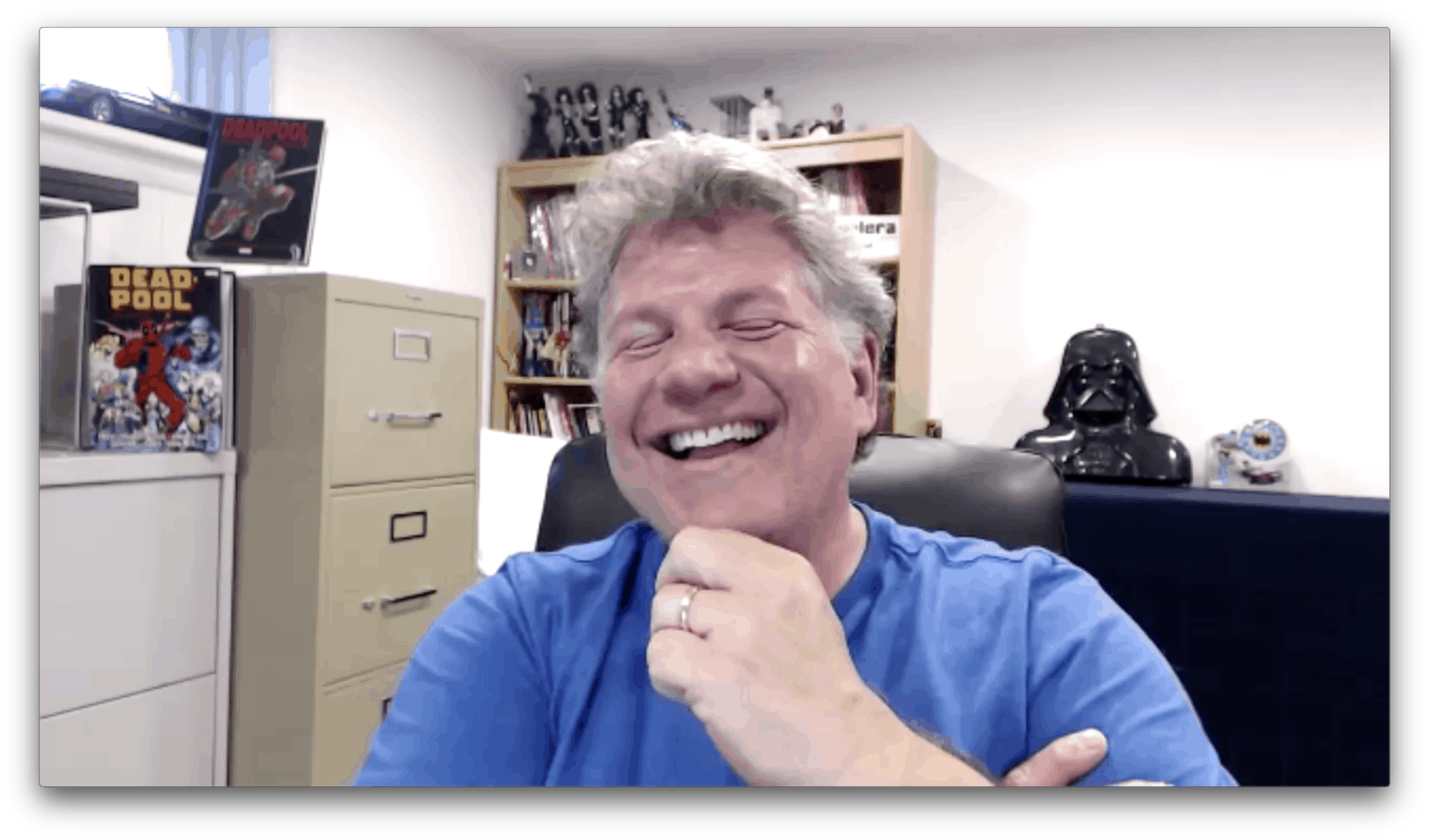 Buddy Scalera riendo en una videollamada, con arte de cómics y figuras de colección al fondo.