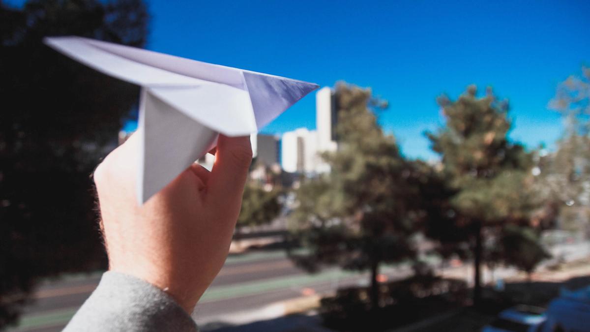 A hand about to throw a paper airplane against a blue sky with trees and buildings in the background